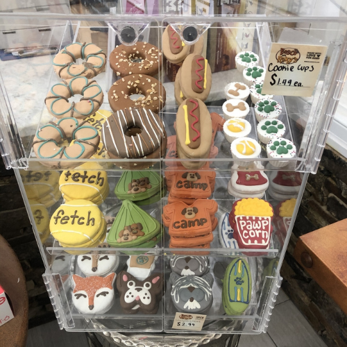 A display case with colorful dog treats shaped like pretzels, hot dogs, and paws. Labeled sections include 