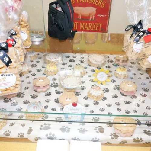 Bakery display with dog-themed treats, including cookies shaped like bones and paw prints. Packaging features black ribbon and paw print motifs. Cozy, inviting atmosphere.