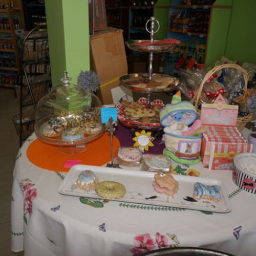 A table in a shop displays assorted baked goods, colorful cookies, and pastries on a floral cloth. The setting is vibrant and inviting.