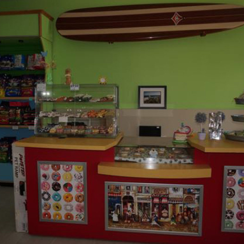 A vibrant bakery counter with assorted pastries in glass displays, colorful doughnut art on the front, and a surfboard mounted on a lime green wall.