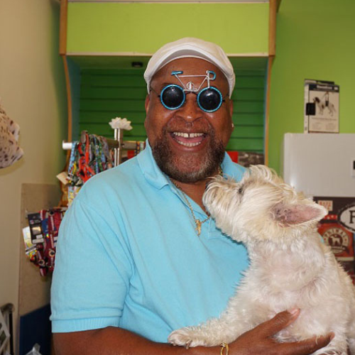 Man in a light blue shirt, wearing humorous glasses and a white cap, joyfully holds a small white dog in his arms inside a colorful room.