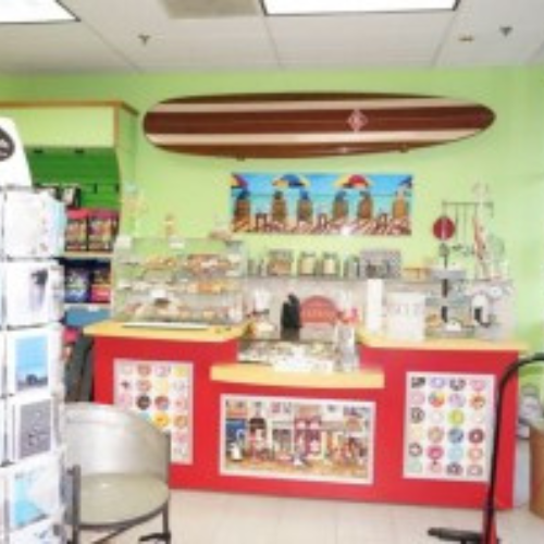 A colorful snack bar with a red counter displaying various candies and snacks. A surfboard decorates the bright green wall above, evoking a cheerful beach vibe.