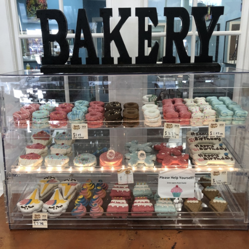 Bakery display case with colorful, decorative dog treats shaped like cupcakes, donuts, and cookies. Prices marked, 'Please Help Yourself!' sign visible.