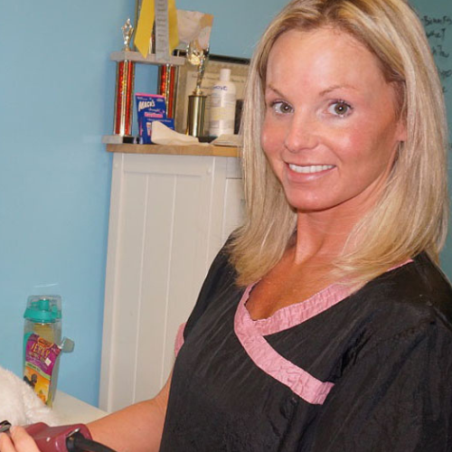 A smiling woman in a black and pink top holding clippers. She stands in a pet grooming salon, with shelves of trophies and grooming supplies behind her.