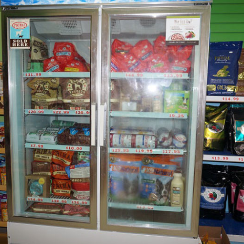 A refrigerated display case with glass doors, filled with various pet food products. Shelves hold bags and cans with visible price tags. Store setting.