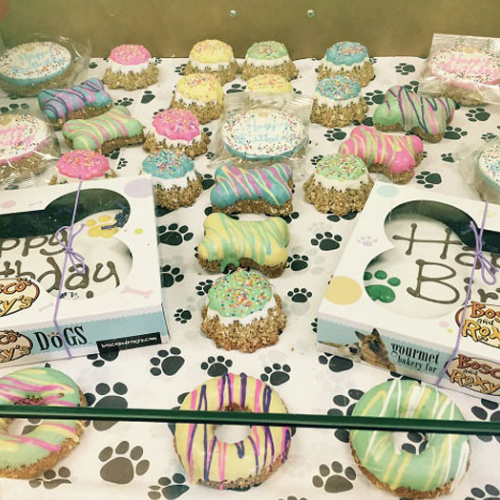 A display of colorful dog treats, including donuts and biscuits, decorated with pastel icing and sprinkles. They rest on paw-print paper. Festive and playful atmosphere.