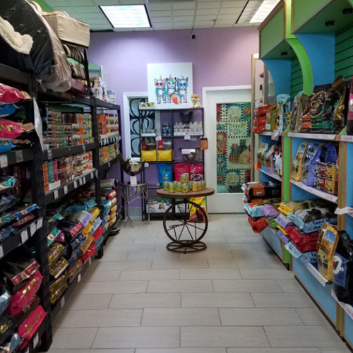 Pet store aisle with shelves filled with colorful bags of pet food and supplies. A round table displays items in the center. Bright, cheerful ambiance.