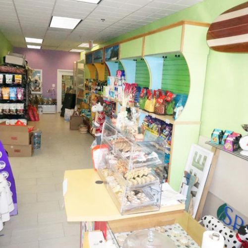 A colorful pet store interior with shelves of pet food, treats, and toys. A glass display case houses various pet snacks near the checkout counter.
