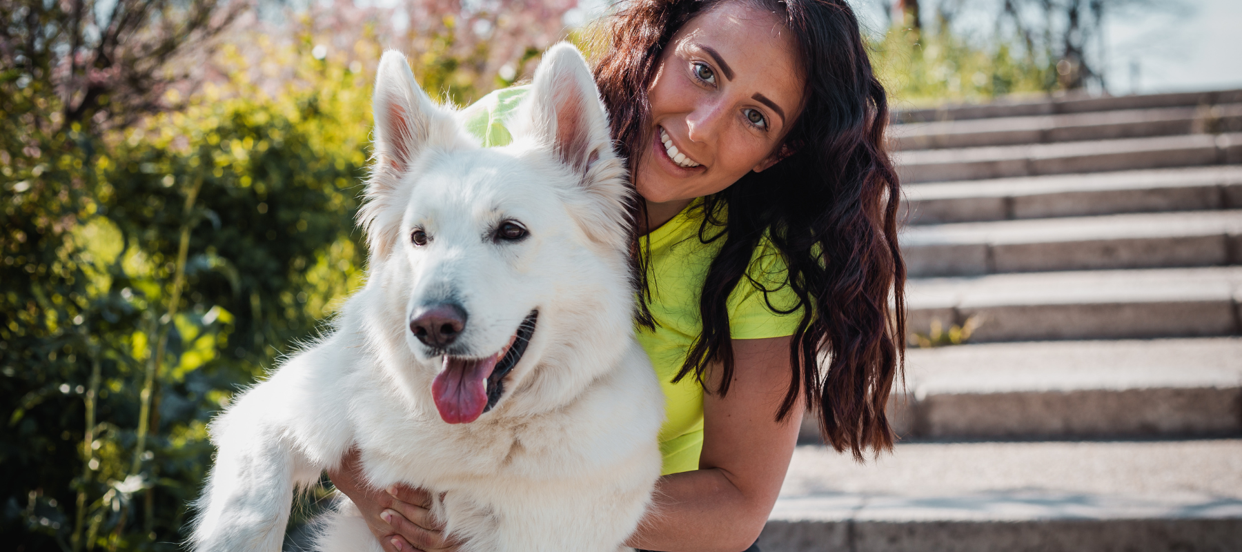 A woman with long dark hair, wearing a yellow shirt, hugs a large white dog on a sunny day. Both are smiling near stone steps and greenery.