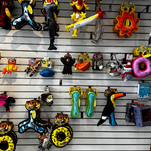 A display wall filled with colorful plush dog toys, including animals and novelty shapes, set in a pet store with a playful and vibrant tone.